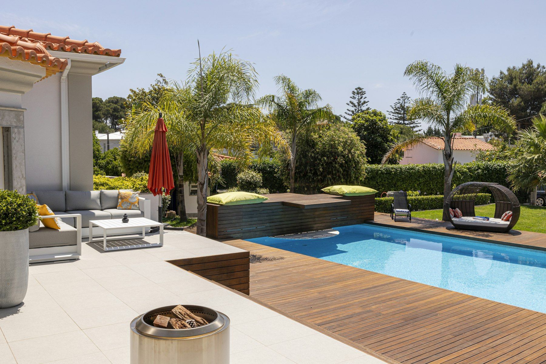 A timber pool deck beside a tiled terrace with outdoor lounges, palms and a fire pit
